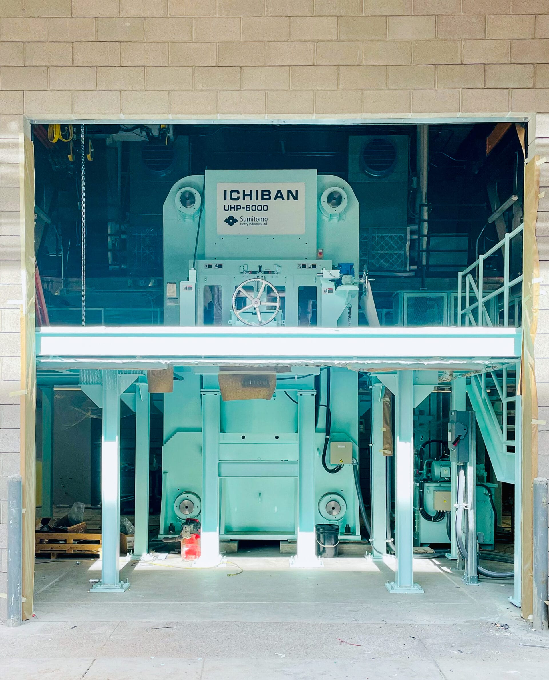 A photograph of a large piece of equipment viewed through a large hole in the side of a building. The piece of equipment is light aqua blue and has a sign on the upper part of the front side that reads, 